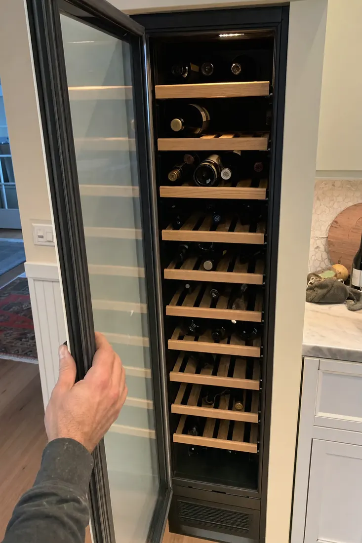 An open built-in wine column, bottles on wooden shelves, a hand at the door seal, the door gasket visible in the picture - Terms of Service